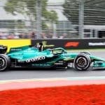 Aston Martin F1 aracı pistte yarışırken, arka planda takım personeli çalışıyor.