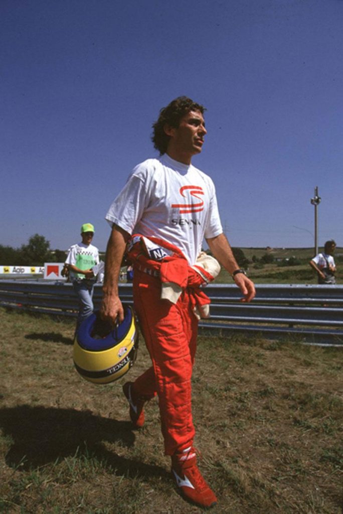 Ayrton Senna kaskini elinde tutarken pist kenarinda yuruyor - kirmizi tulum Senna branded tisort, Nacional kask