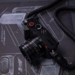 Leica Q3 43 Formula 1 Edition — F1 araç tasarım eskizleri üzerinde kamera, kırmızı deklanşör butonu, karbon fiber başparmak desteği ve örgülü F1 kayışı
