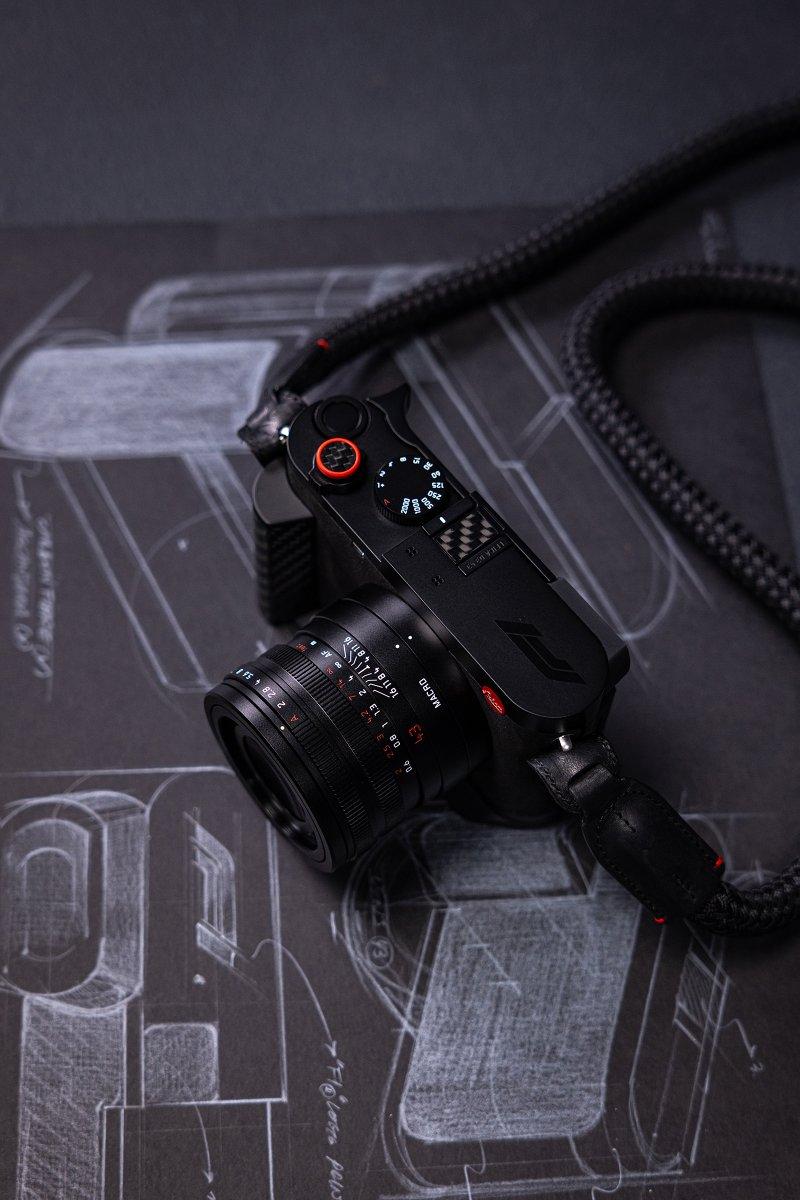 Leica Q3 43 Formula 1 Edition — F1 araç tasarım eskizleri üzerinde kamera, kırmızı deklanşör butonu, karbon fiber başparmak desteği ve örgülü F1 kayışı